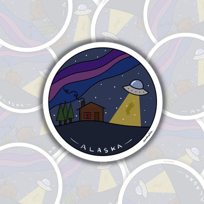 on the left of the sticker are green trees, in the middle a brown cabin with a line of smoke coming out of the chimney. on the right is a ufo with a light beam coming out the bottom. in the light beam is a bear being abducted. in the background are white dots for stars and northern lights stretching from the left to the right of the sticker.