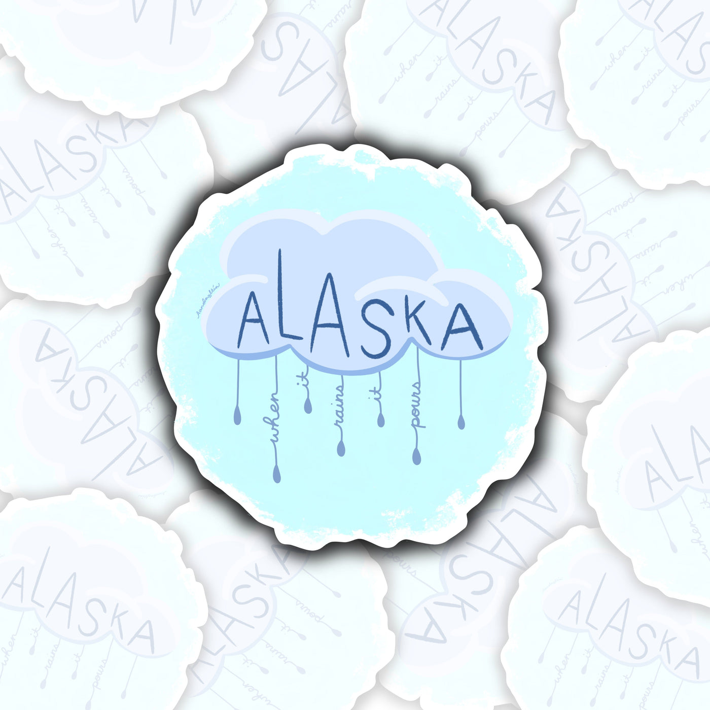 in the upper center of the sticker is a blue-grey cloud. in the cloud it says alaska. coming from the bottom of the cloud are lines of rain. in the lines, it says in cursive "when it rains it pours"